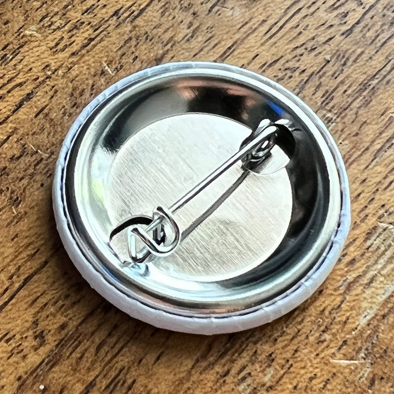 Back of a round white button with a shiny silver metal pin clasp, on a textured wooden surface.