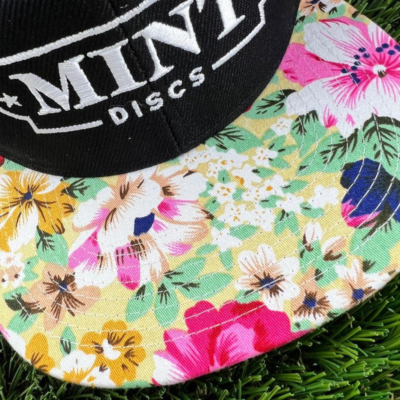 Black mint discs snapback hat with a vibrant yellow, pink, and green floral patterned flat bill on green turf.