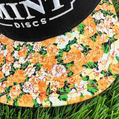 Black and white "mint discs" logo on a black snapback hat, with an orange, yellow, and green floral flat bill resting on.
