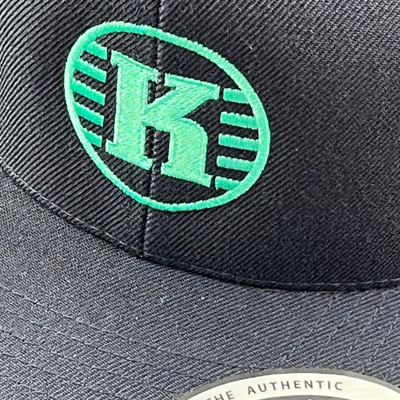 A black yupoong kastaplast snapback hat with a vibrant green embroidered "k" logo and an oval outline.