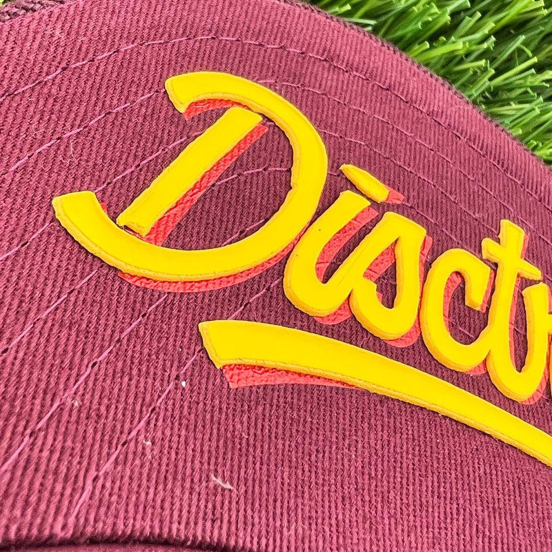 Close-up of a burgundy disctroyer flat bill snapback hat with thick yellow "disctroyer" lettering.