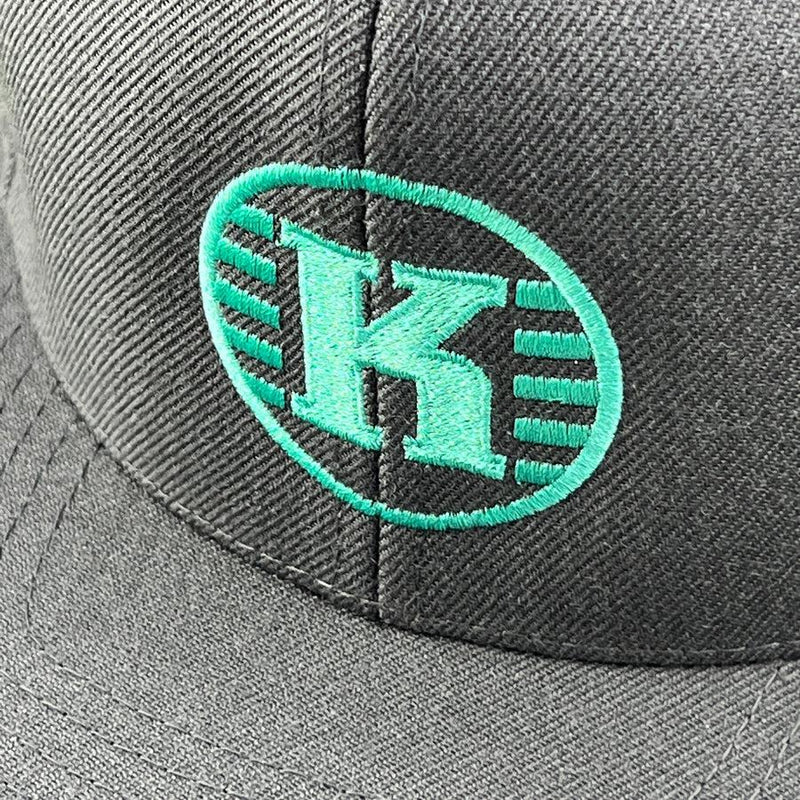 A close-up of a charcoal gray yupoong hat with a bright teal embroidered kastaplast "k" logo.
