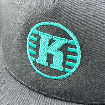 Close-up of a charcoal grey yupoong kastaplast hat with an embroidered bright teal "k" inside a circle logo.