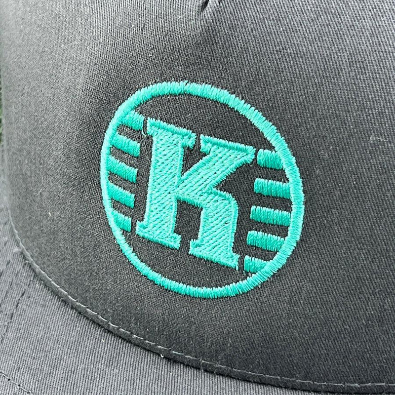Close-up of a charcoal grey yupoong kastaplast hat with an embroidered bright teal "k" inside a circle logo.