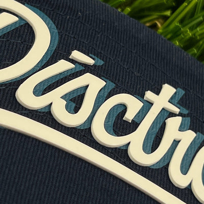 Close-up of a dark blue canvas disctroyer hat with raised white and teal lettering.
