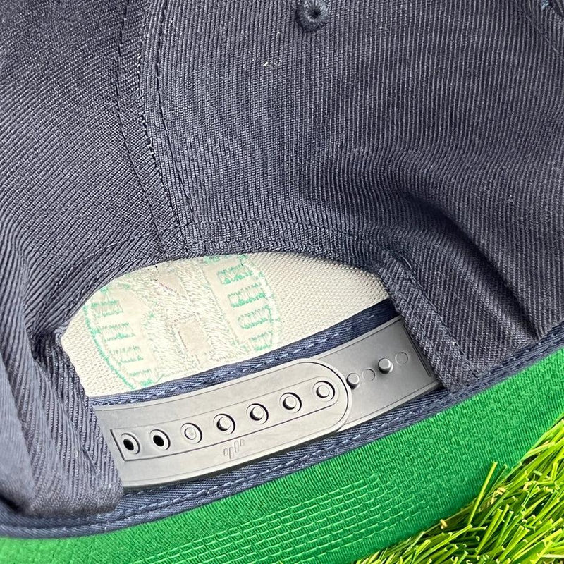 Close-up of a dark navy yupoong kastaplast snapback hat from the back, showing its grey plastic closure and green brim,.