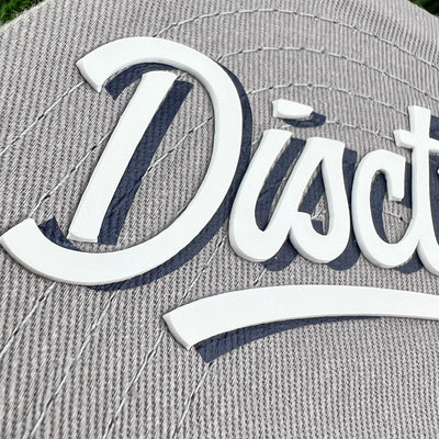 Close up of discstroyer with white lettering and navy blue shadow on a grey ribbed flat bill snapback hat.