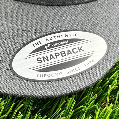 A close-up of a gray yupoong snapback clothback hat's brand sticker resting on green artificial turf.