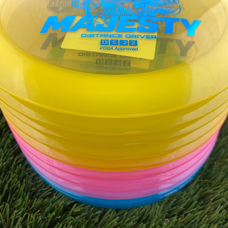 Stacked yellow, pink, and blue Discmania Majesty Distance Driver disc golf discs with blue lettering and flight numbers.