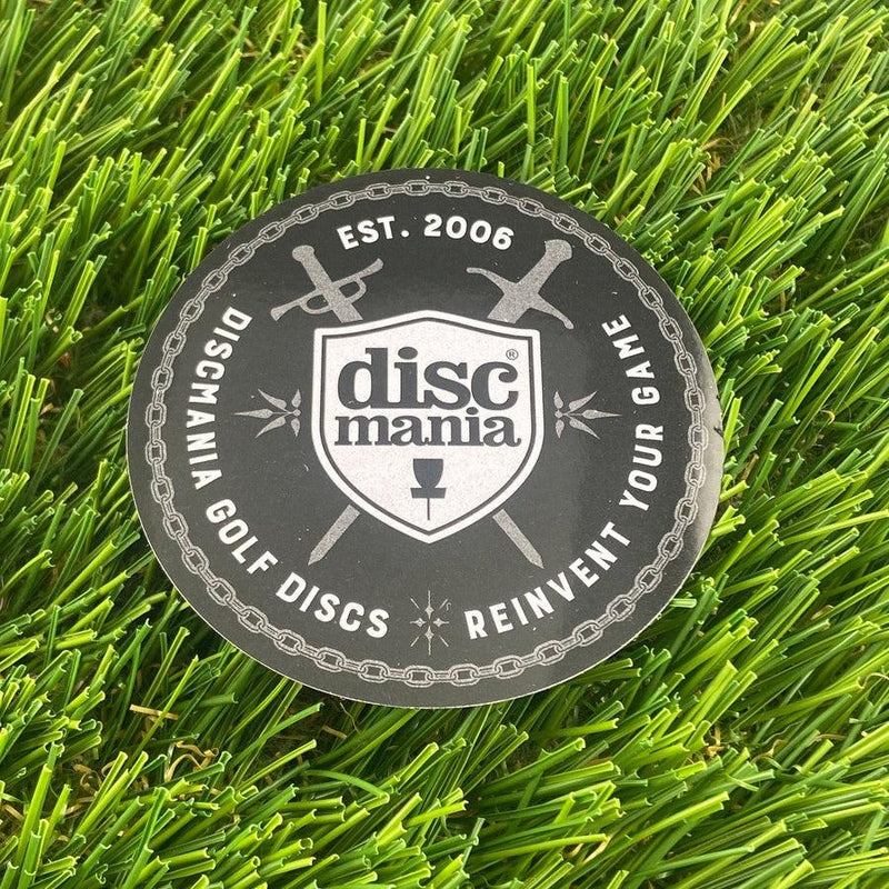 Black Discmania circular sticker with a white shield logo, crossed swords, and the text "REINVENT YOUR GAME".