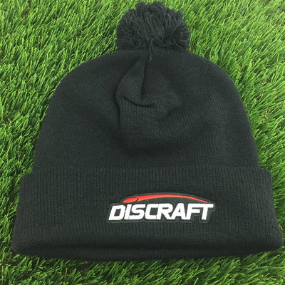 Black knit Discraft embroidered logo cuff pom beanie with a red stripe above white lettering.