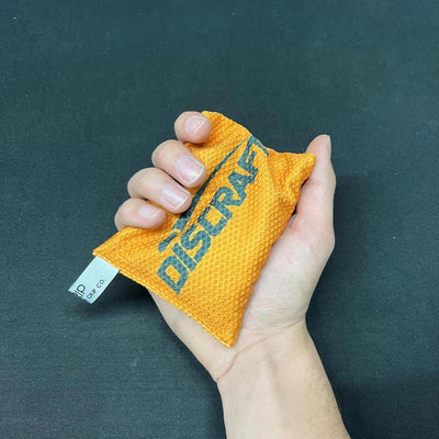 Person's hand gripping an orange Discraft Silky Feel Waffle Sportsack with a black Discraft logo and a white tag.