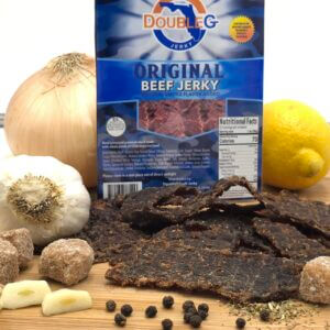 Double G Original Beef Jerky packaging stands behind a pile of dark brown jerky pieces, with garlic, onion, and lemon nearby.