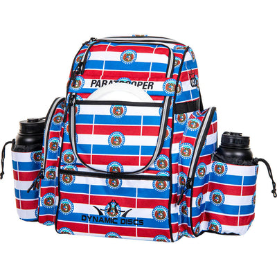 Colorful Dynamic Discs Paratrooper backpack disc golf bag with red, white, and blue stripes and two water bottles.