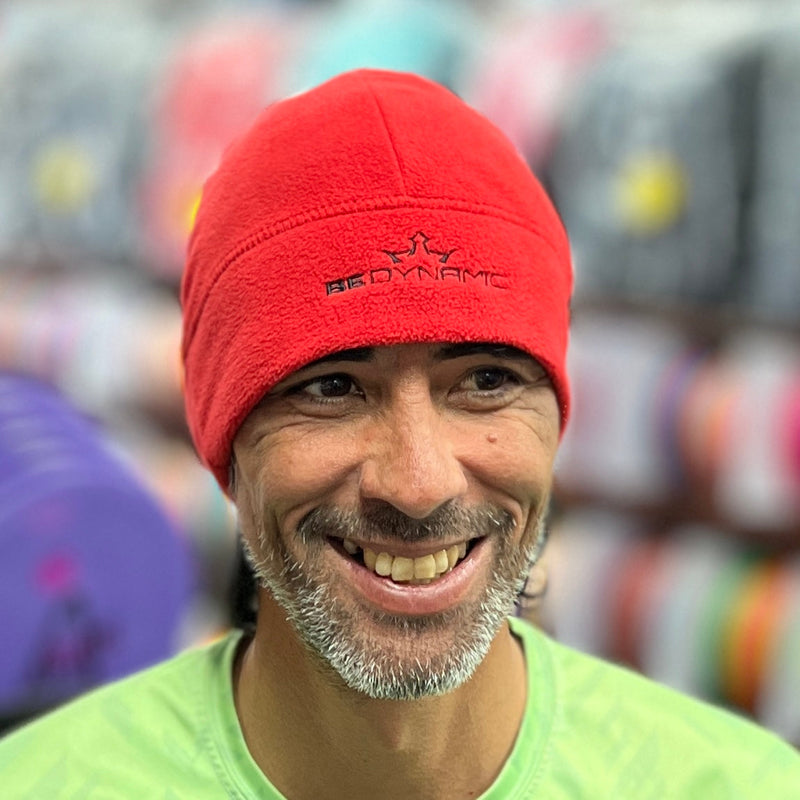 Smiling man wearing a red fleece beanie with a black embroidered "BEDYNAMIC" logo and crown design.