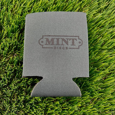 Gray neoprene beverage koozie featuring the "mint discs" logo, resting on vibrant green artificial turf.