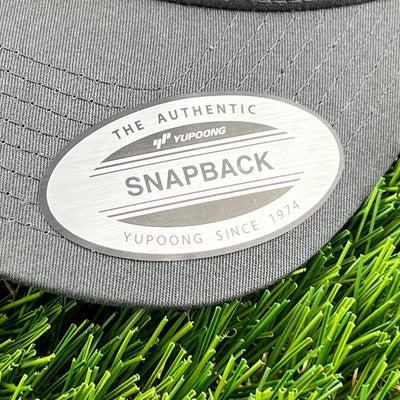 A gray yupoong the authentic snapback hat label on a green faux grass background with the hat out of view.