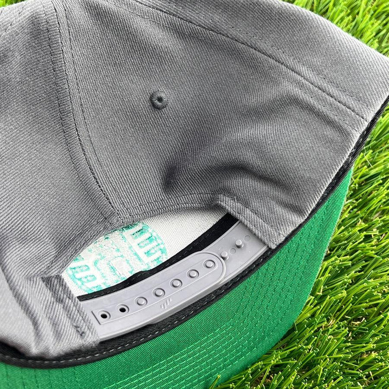 Gray yupoong kastaplast snapback hat with a green brim and a gray plastic snap closure, resting on grass.