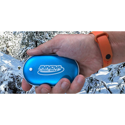 A person's hand holds a blue Innova Disc Golf rechargeable pocket warmer with a white button.