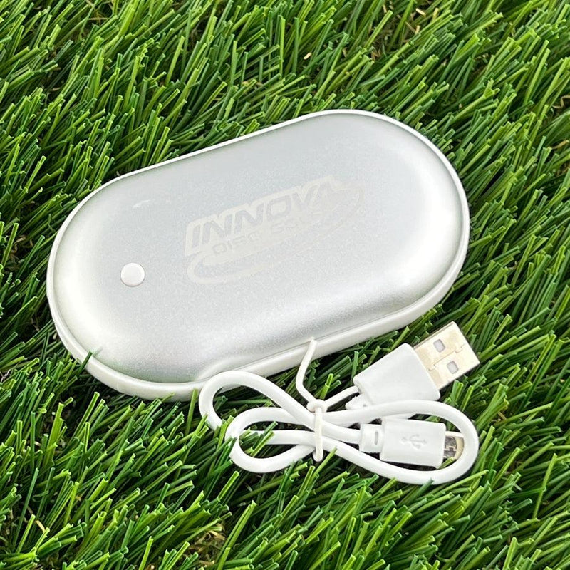 Silver metallic Innova rechargeable hand warmer with a small white indicator light and a coiled white USB charging cable.