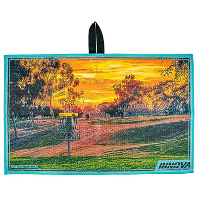 Innova Tour Towel featuring a vibrant sunset disc golf course with a basket and yellow flag, bordered in teal.