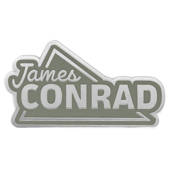Flat gray James Conrad enamel pin with a silver metallic border and bold lettering.