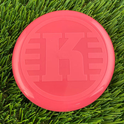 Round, bright salmon pink Kastaplast K Mini Marker disc with an embossed capital K and horizontal lines.