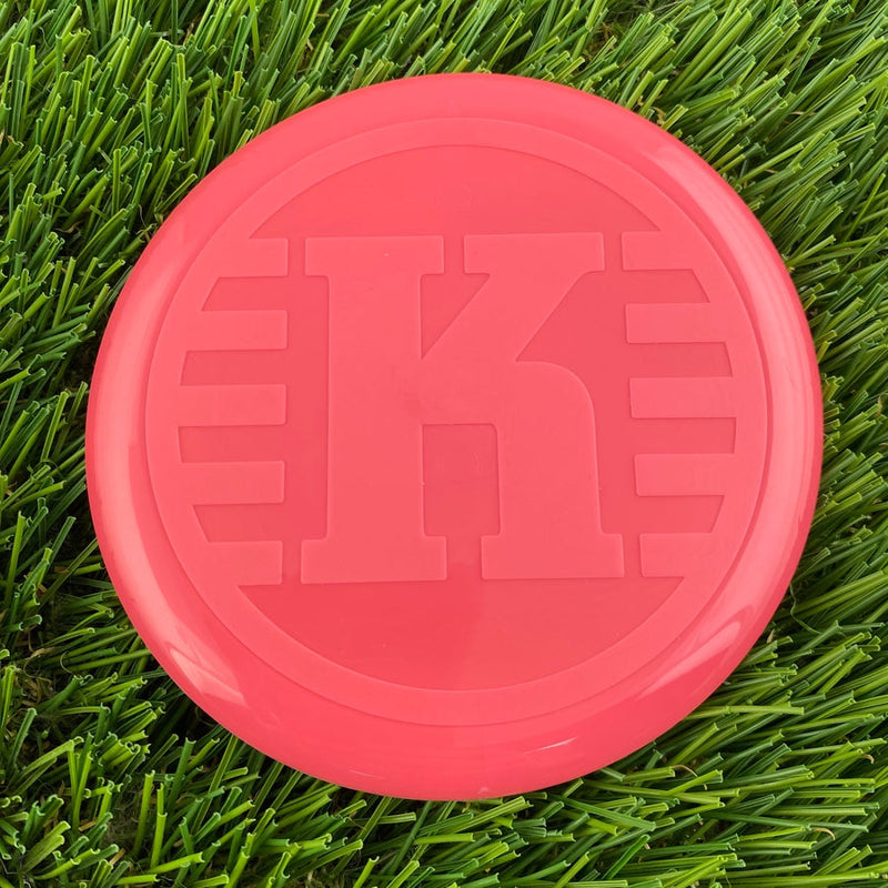Round, bright salmon pink Kastaplast K Mini Marker disc with an embossed capital K and horizontal lines.