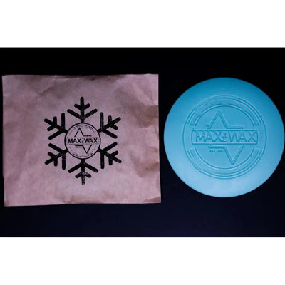 A light blue circular max wax mini grip wax for disc golf beside its brown paper packaging with a snowflake logo.