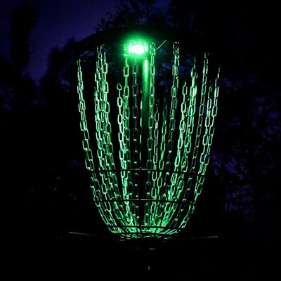 A metallic disc golf basket with shiny chrome chains glowing brightly with green light in a dark setting.