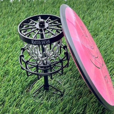 A miniature MVP Black Hole Micro disc golf basket with silver chains stands next to a pink MVP disc.