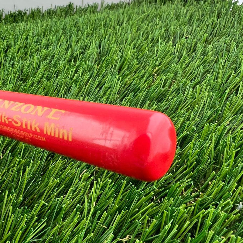 A red "inzone kwik-stik mini" pole with yellow lettering lies diagonally on artificial green grass.