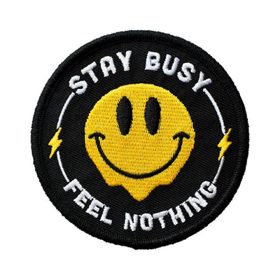 Black round iron-on patch with a yellow melting smiley face and white embroidered text that says "STAY BUSY FEEL NOTHING".