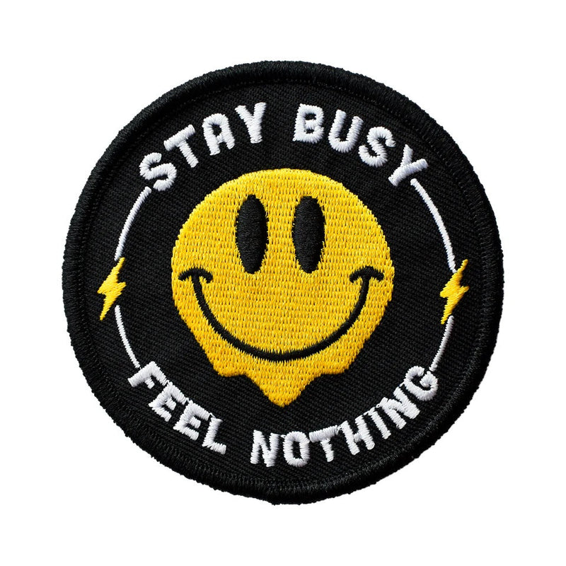 Black round iron-on patch with a yellow melting smiley face and white embroidered text that says "STAY BUSY FEEL NOTHING".