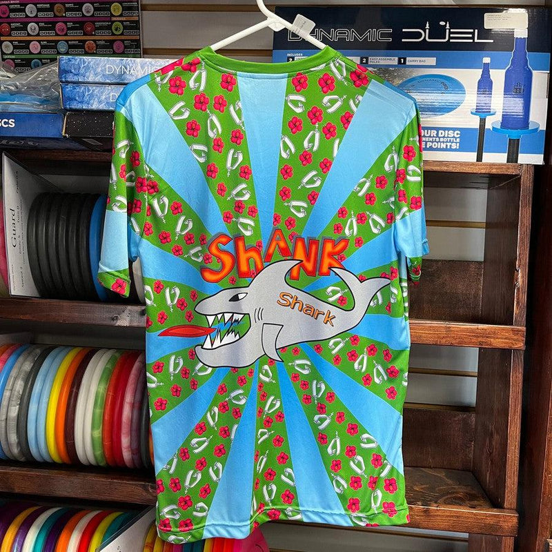 Vibrant Shank Shark printed jersey featuring a gray shark, "Shank" lettering, green background, blue rays, and red flowers.