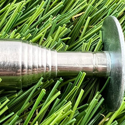 A shiny, silver inzone kwik stik mini round hook, featuring a ribbed barrel and a flat head, rests on artificial green grass.