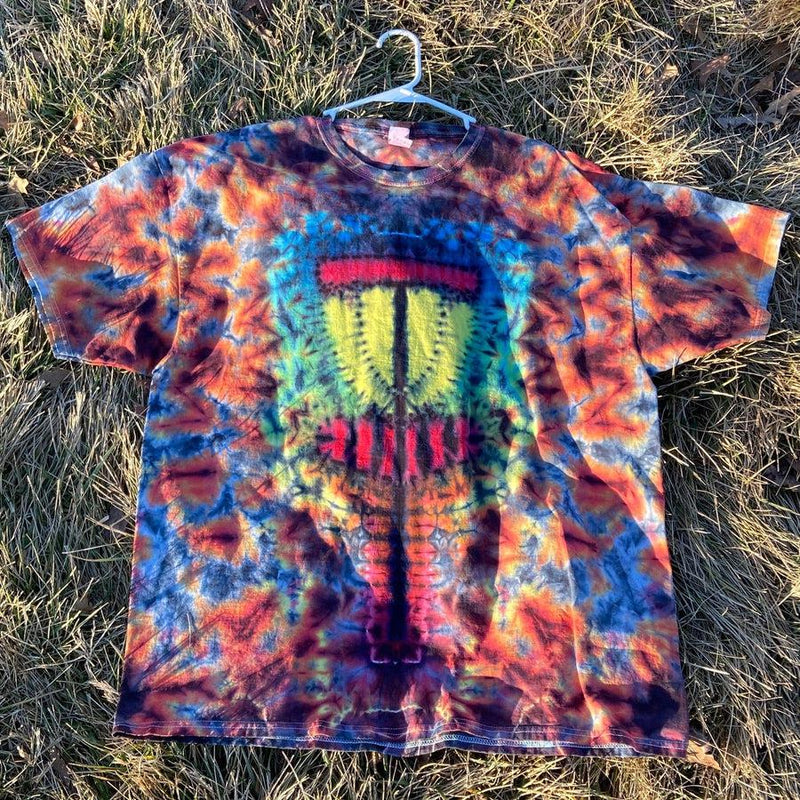 A vibrant tie-dye Twisted Amanita basket-style t-shirt with a detailed yellow, red, and blue disc golf basket graphic.